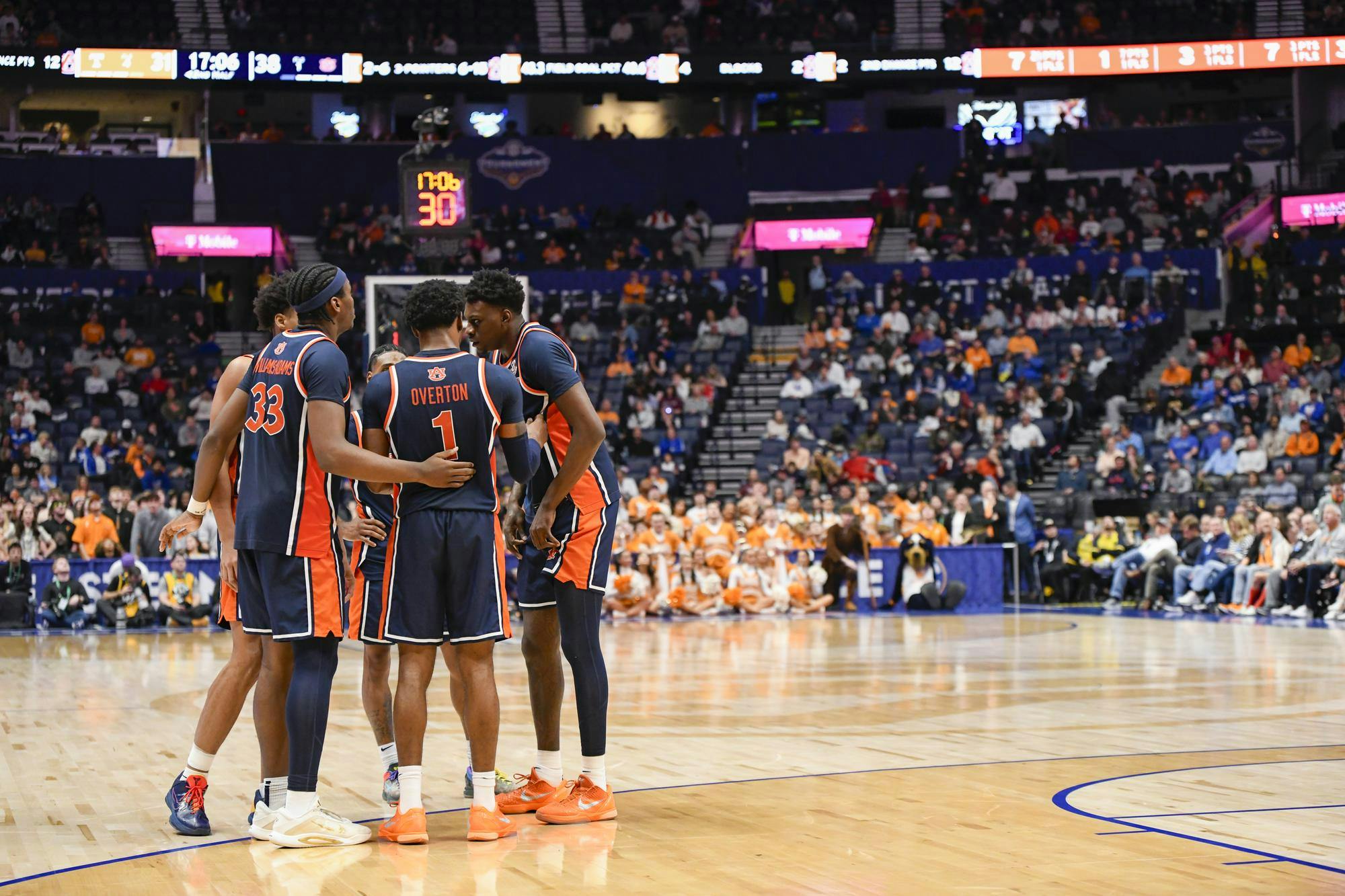 A group of basketball players huddle together on the court with spectators in orange clothing visible in the background.