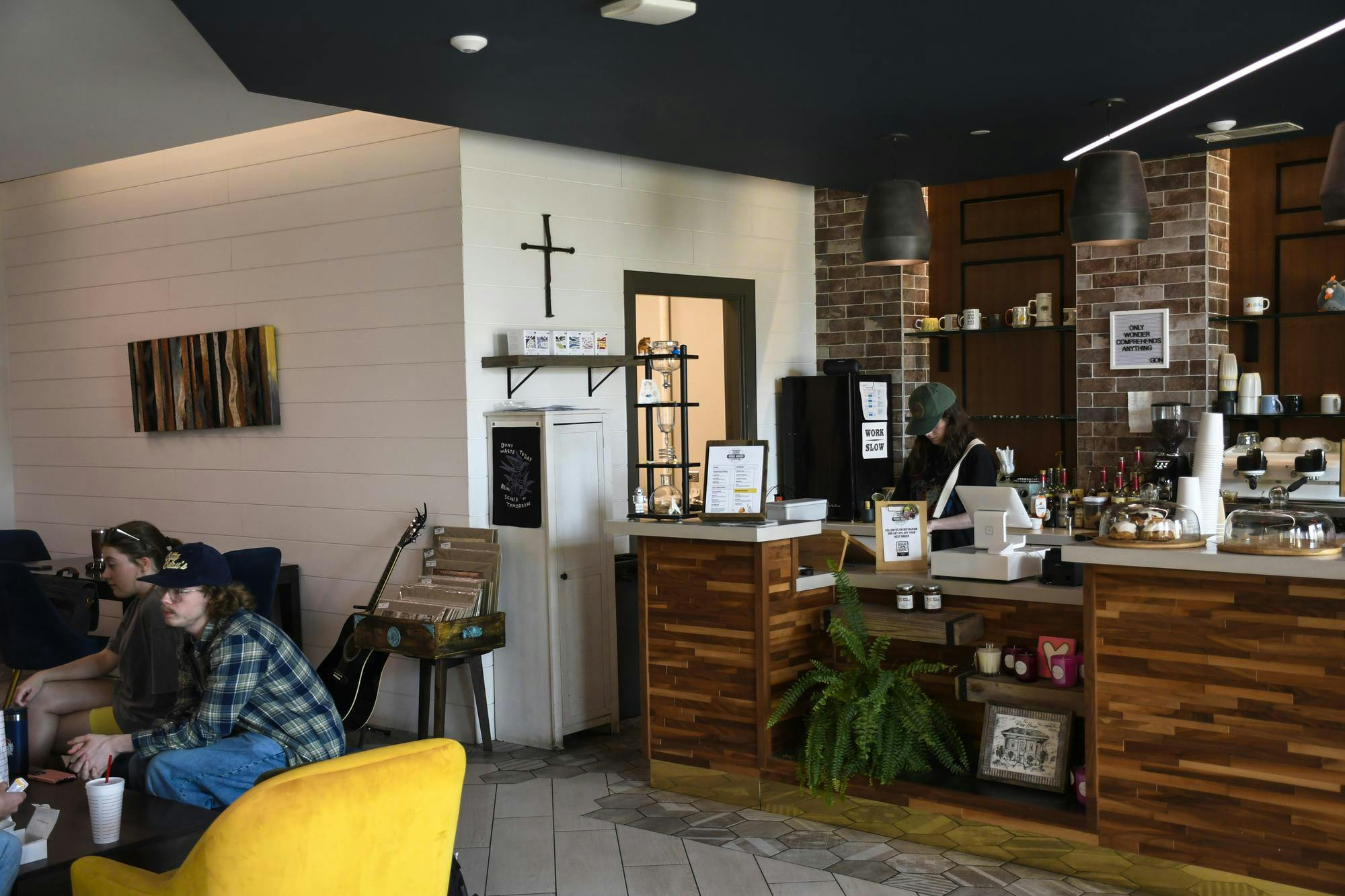 A cozy café features a wooden counter, shelves of mugs, seating areas, and patrons engaged in conversation.