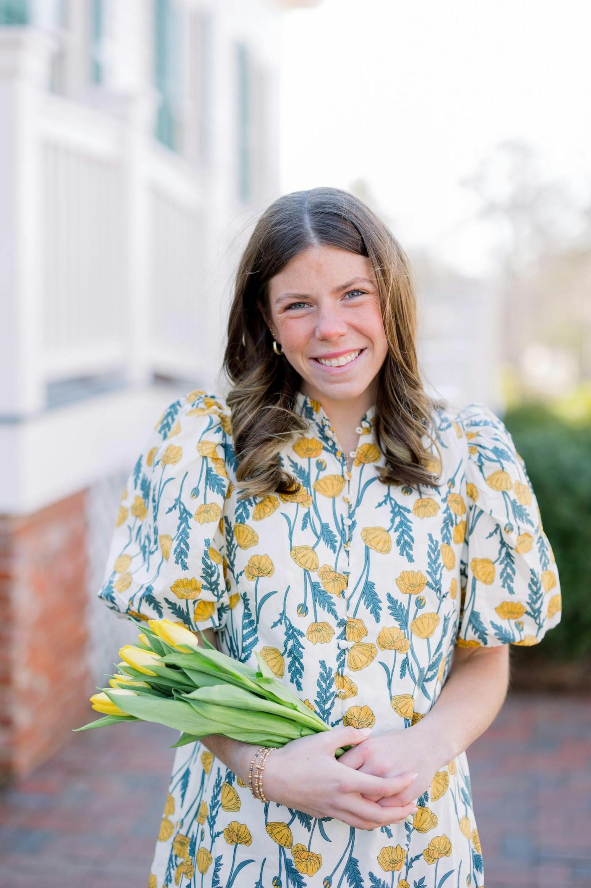 A smiling young woman wears a floral dress and holds a bouquet of yellow tulips. 