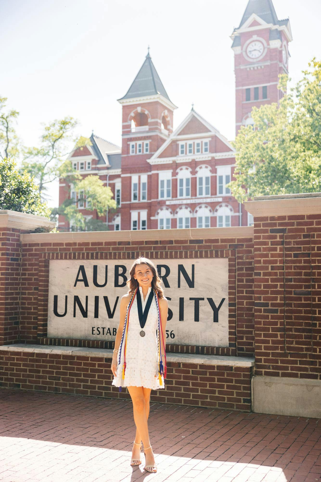 A young woman in a white dress stands in front of an Auburn University sign, wearing multiple academic honor cords and a medal. Contributed by Ally Northridge.