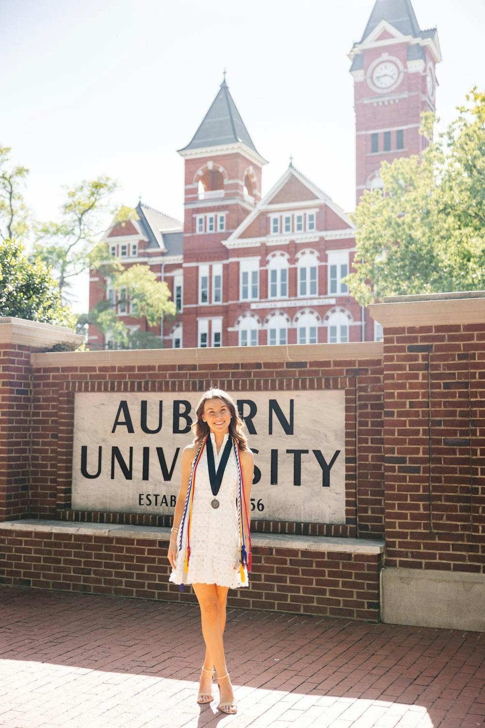 <p>A young woman in a white dress stands in front of an Auburn University sign, wearing multiple academic honor cords and a medal. Contributed by Ally Northridge.</p>