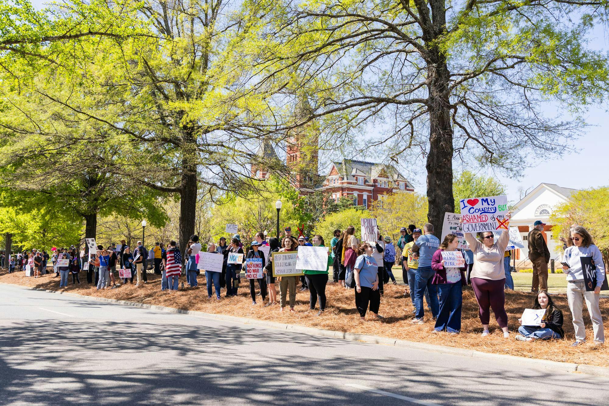 A crowd of people holds protest signs along a tree-lined street, with a historic building visible in the background.