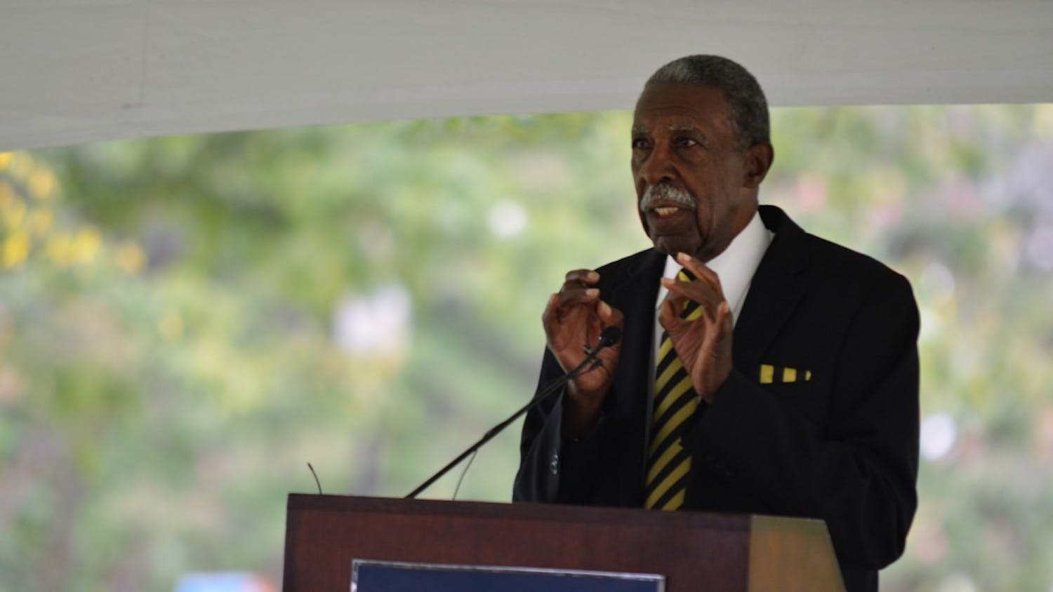 An elderly man in a suit stands at a podium, gesturing while speaking to an audience outdoors.
