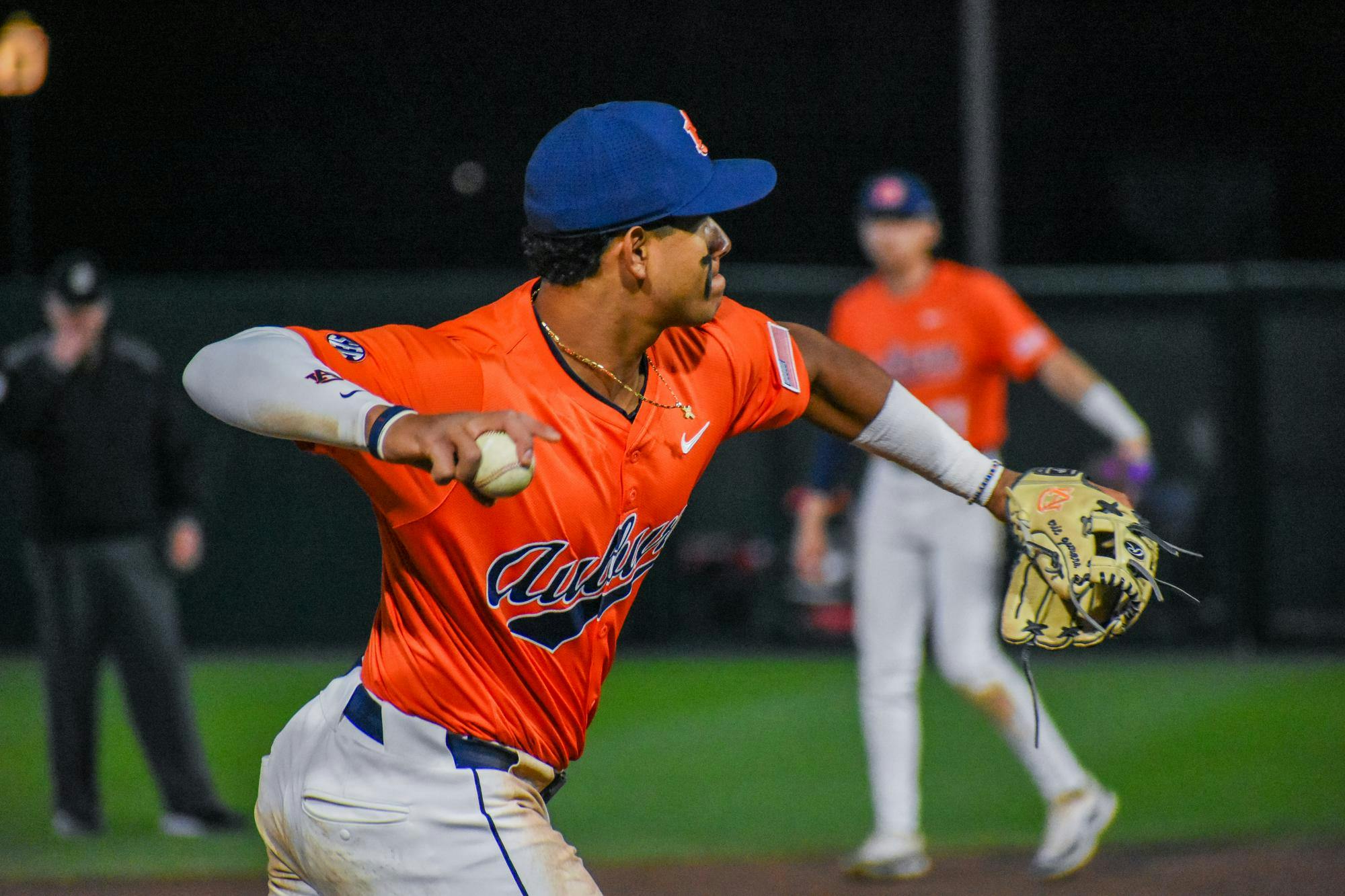A player dressed in an orange jersey and white pants throws a baseball, while another player stands behind him.