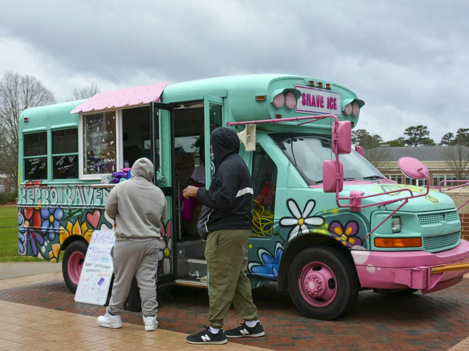 A brightly colored food truck with flowers painted on it is approached by two people in casual attire.