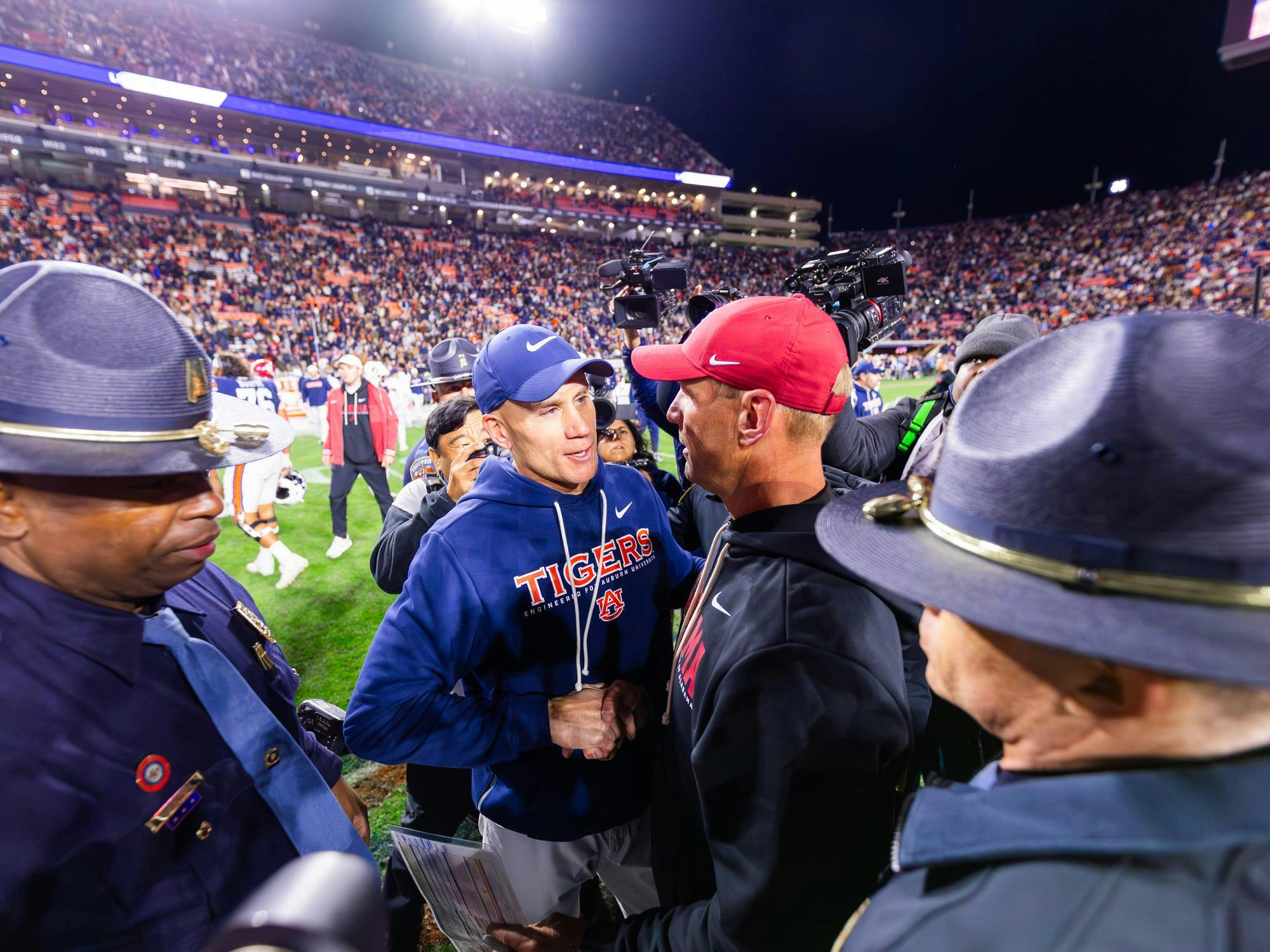 Two coaches in team apparel shake hands amidst a crowd, with police and cameras nearby at a sports event.