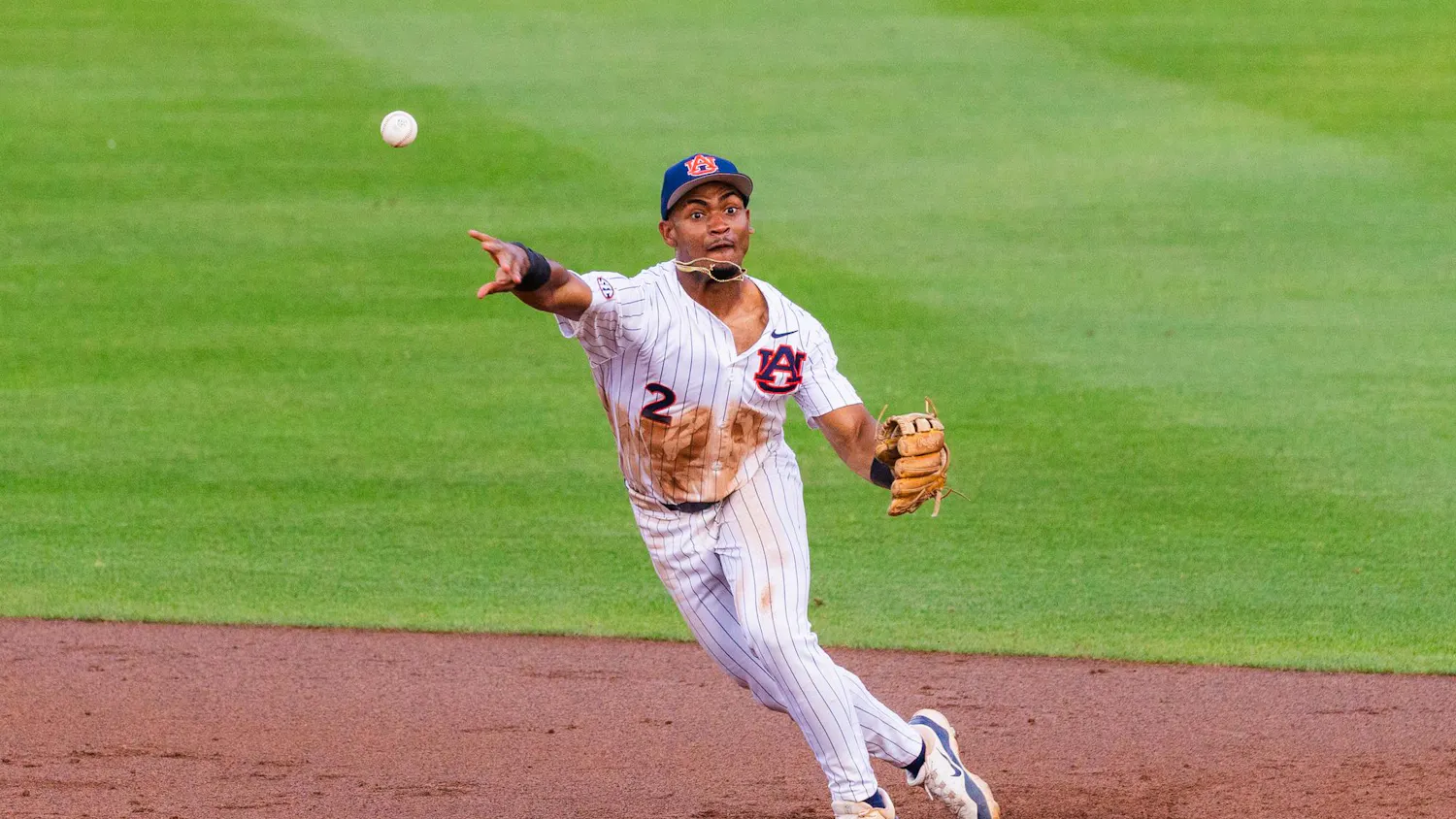 A player in a white and blue striped uniform is throwing a baseball while running on a dirt infield.