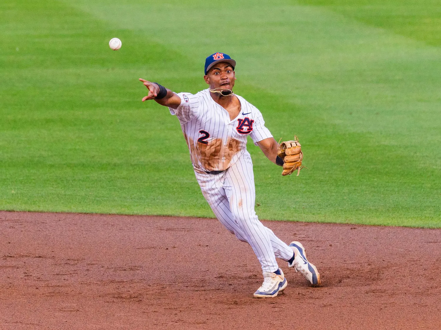 A player in a white and blue striped uniform is throwing a baseball while running on a dirt infield.