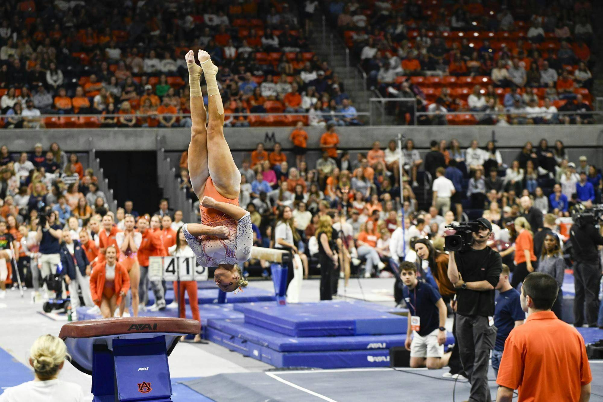A gymnast performs a skill on a vault while surrounded by a cheering crowd and cameras in a sports arena.
