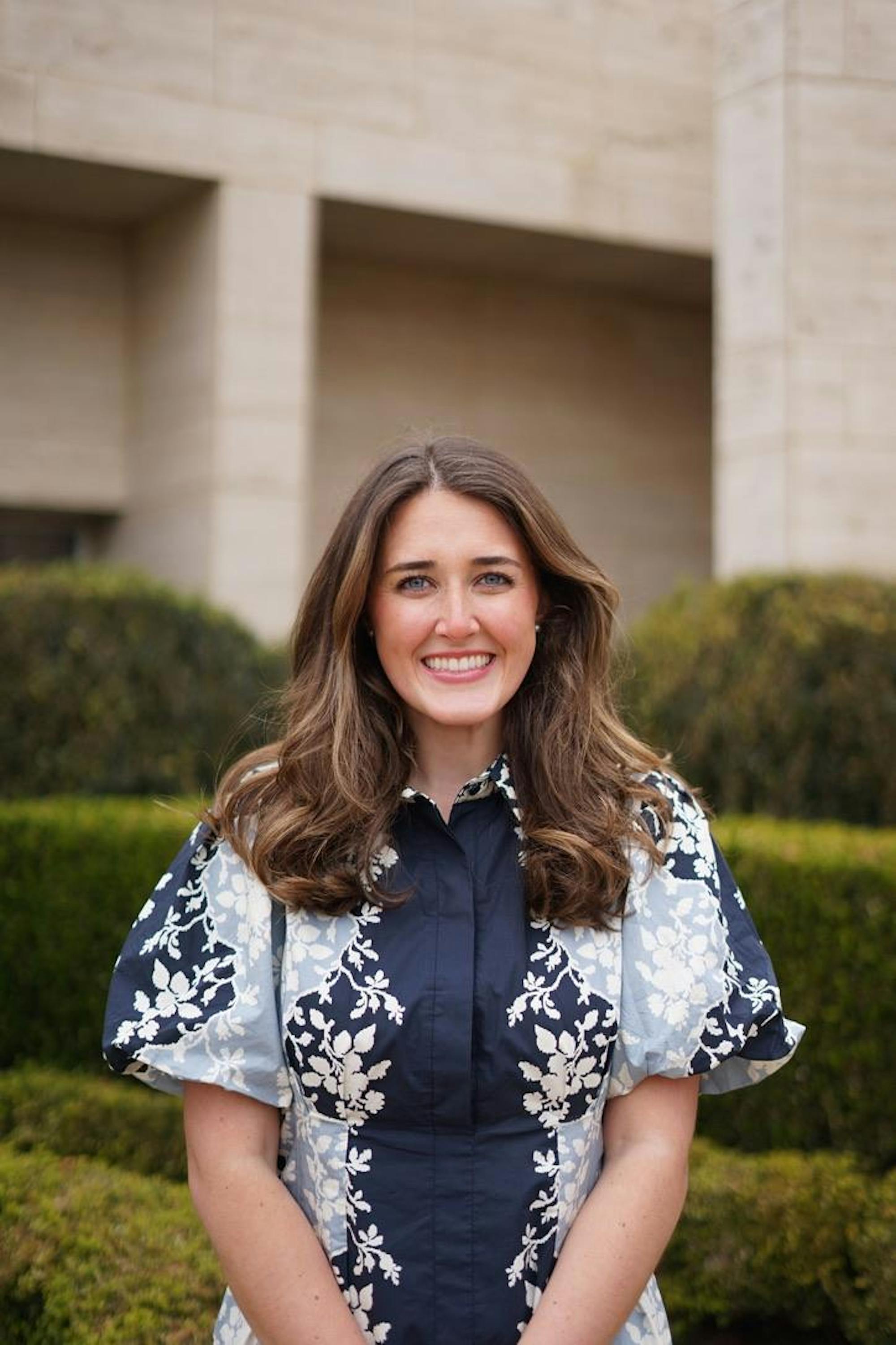 A woman with long, wavy brown hair smiles while wearing a patterned blouse, standing in front of green bushes.