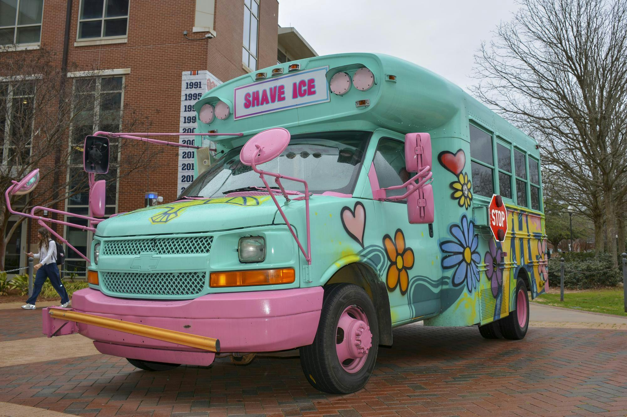 A brightly painted food truck resembling a school bus, adorned with colorful flowers, features a sign that reads "SHAVE ICE."