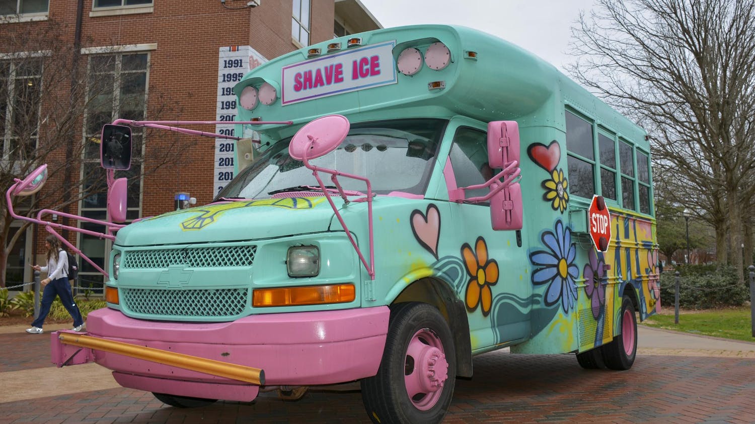 A brightly painted food truck resembling a school bus, adorned with colorful flowers, features a sign that reads "SHAVE ICE."