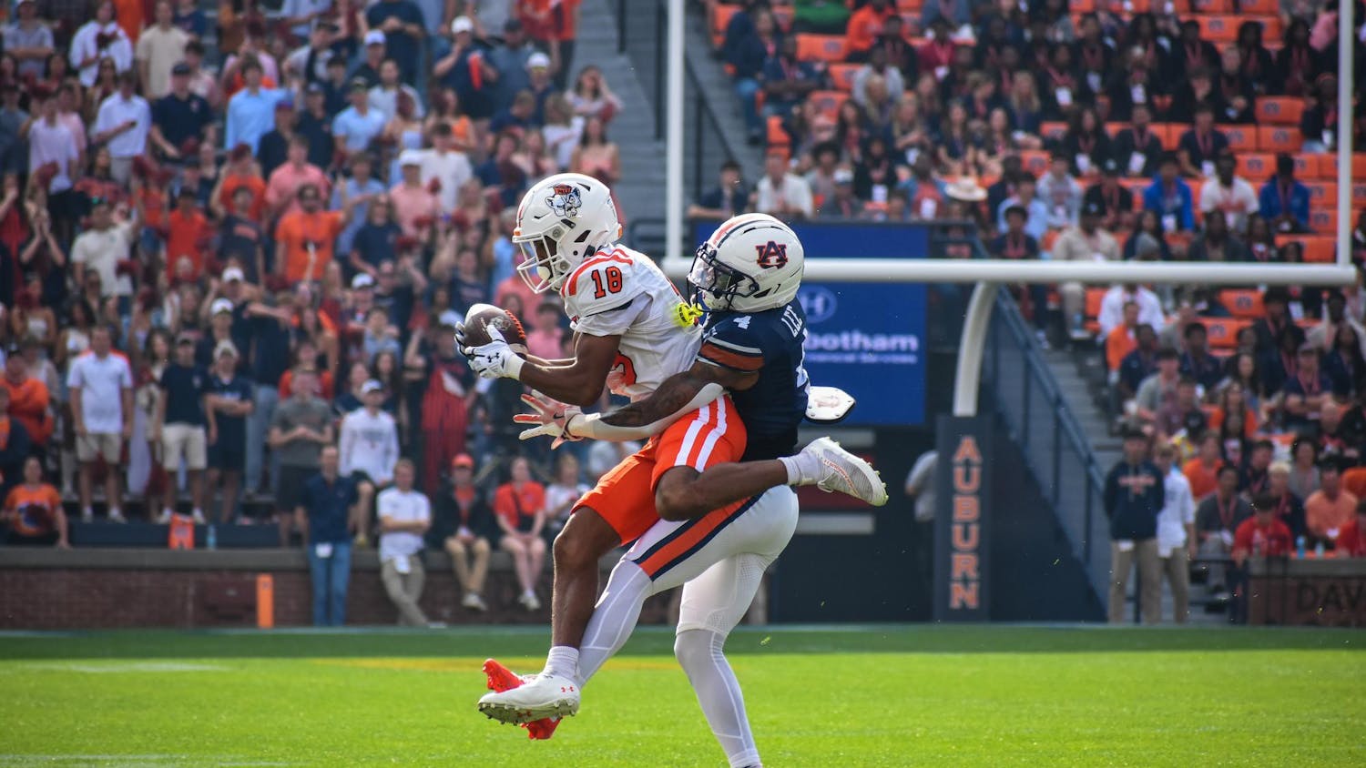 A football player in a white jersey catches a ball while being tackled by another player in a dark jersey on a grassy field.