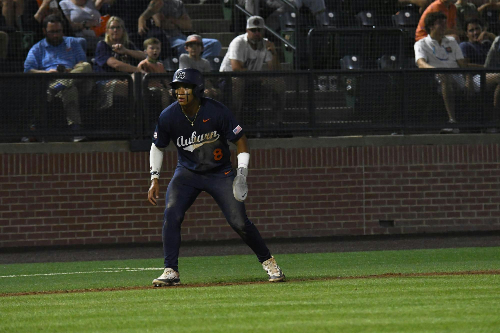 A baseball player in a dark jersey stands on a diamond, preparing to run, with spectators in the background.