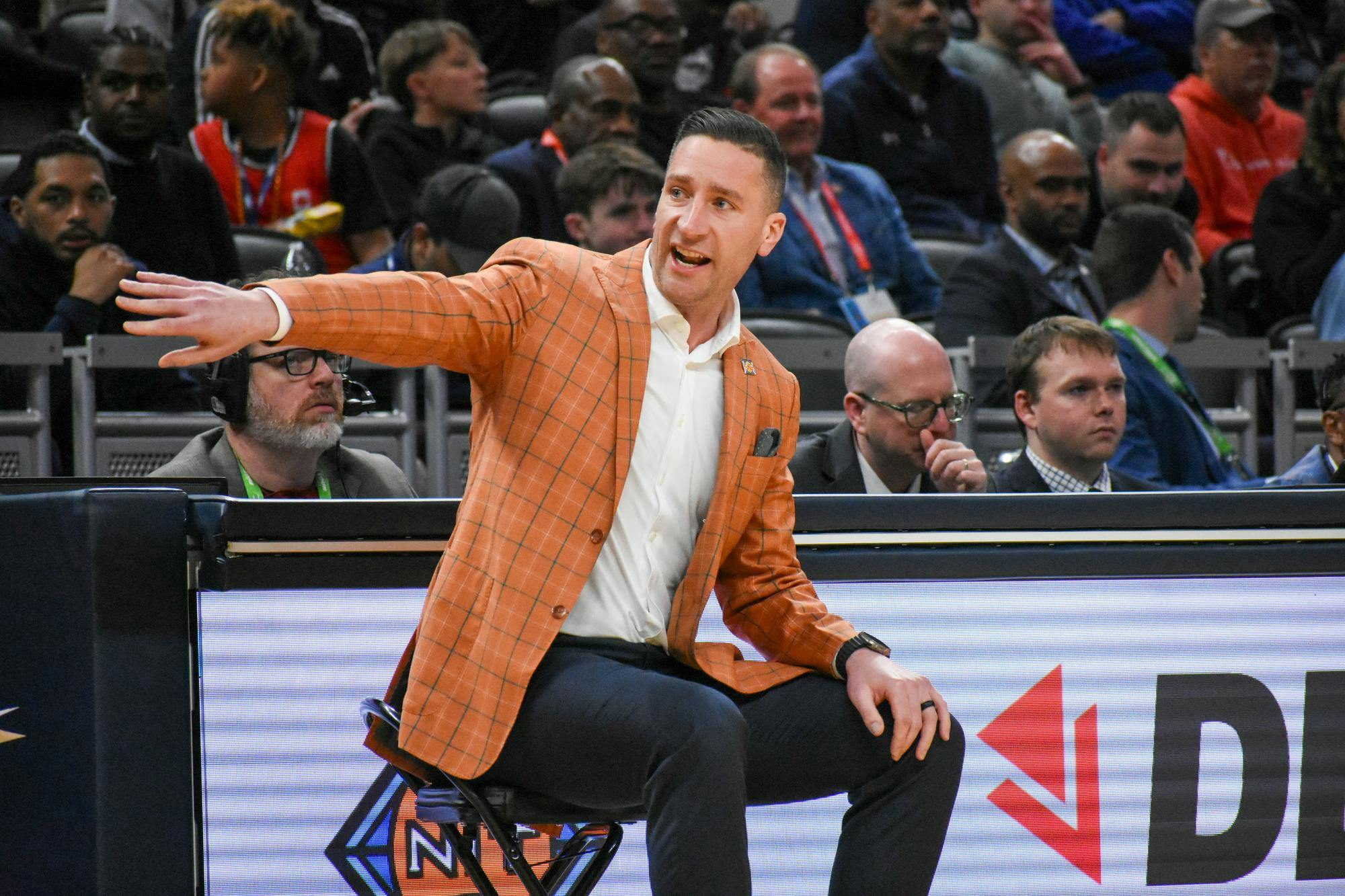 A man in an orange checkered blazer sits on a stool, animatedly gesturing while surrounded by focused spectators.