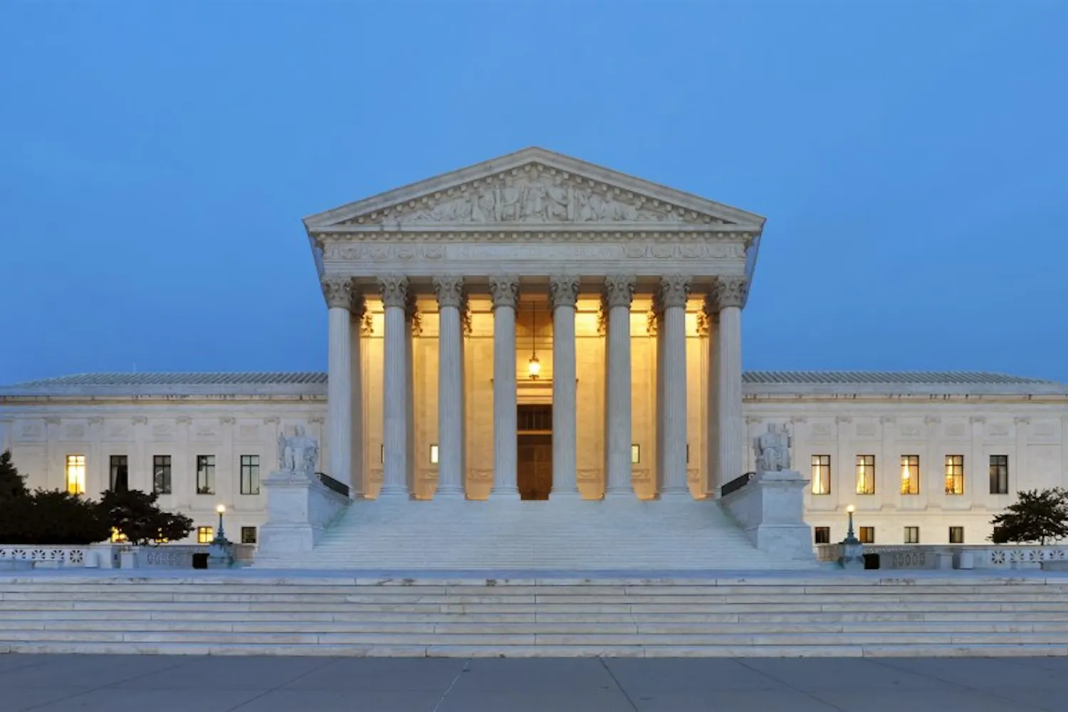 A large, classical building features a series of tall columns, wide steps, and illuminated entrances, set against a twilight sky.