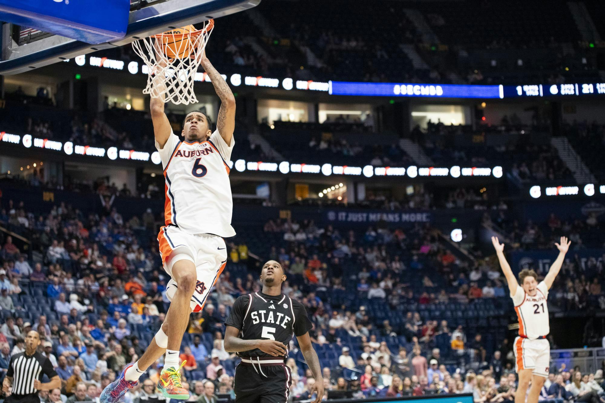 A basketball player in an orange and white uniform jumps to dunk a ball as two teammates react on the court.