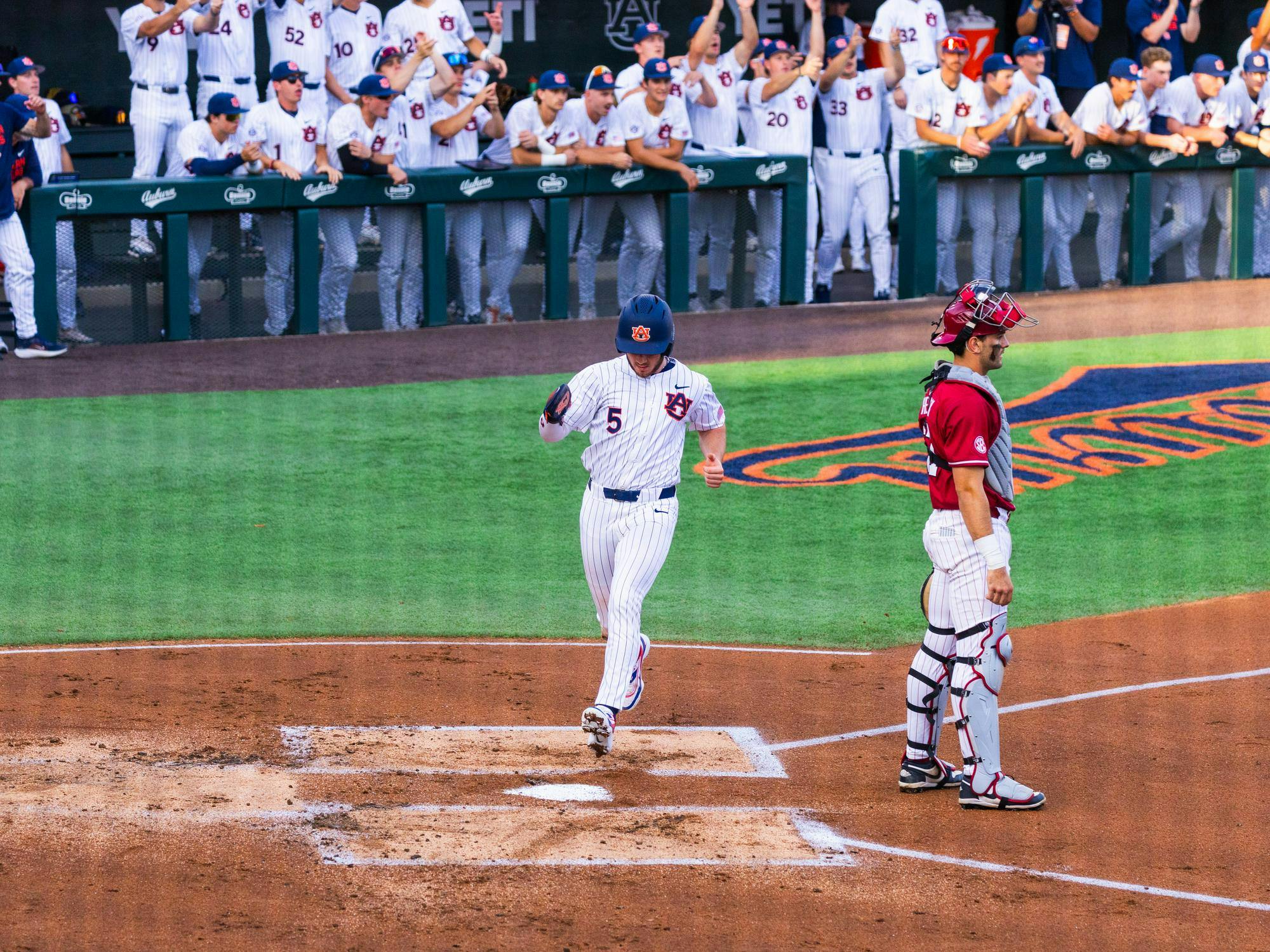 A baseball player in a blue hat and striped uniform crosses home plate, while a catcher in red stands nearby and spectators cheer.