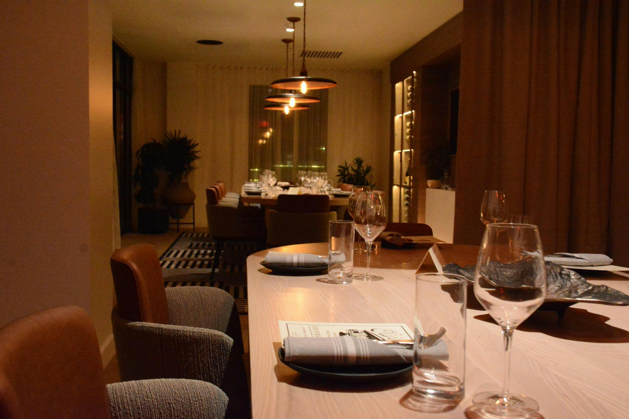 A modern dining area features a long table set with glasses and plates, illuminated by hanging pendant lights, with plants in the background.