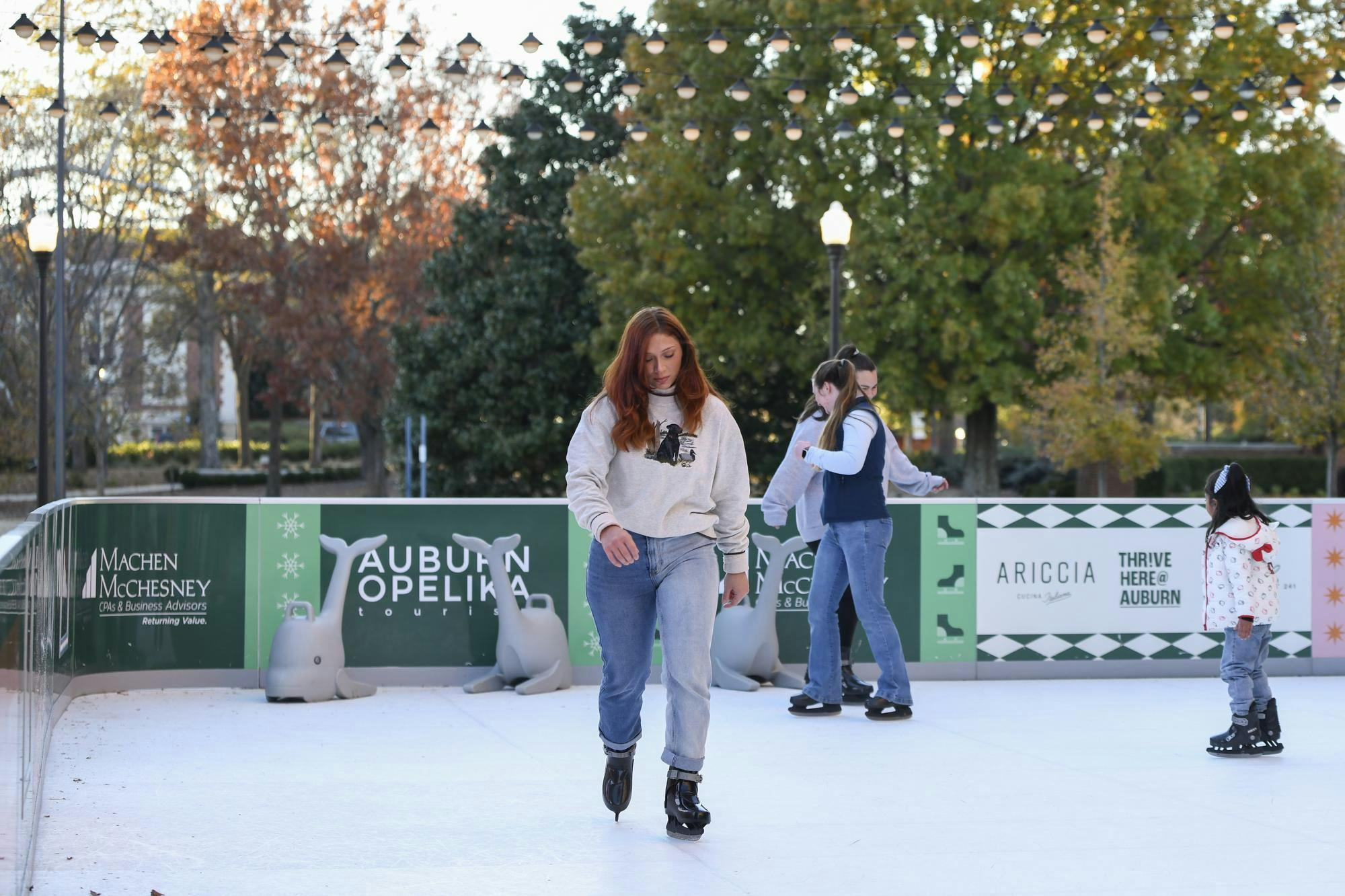 A group of people ice skating on a rink surrounded by green advertisements and colorful trees in the background.