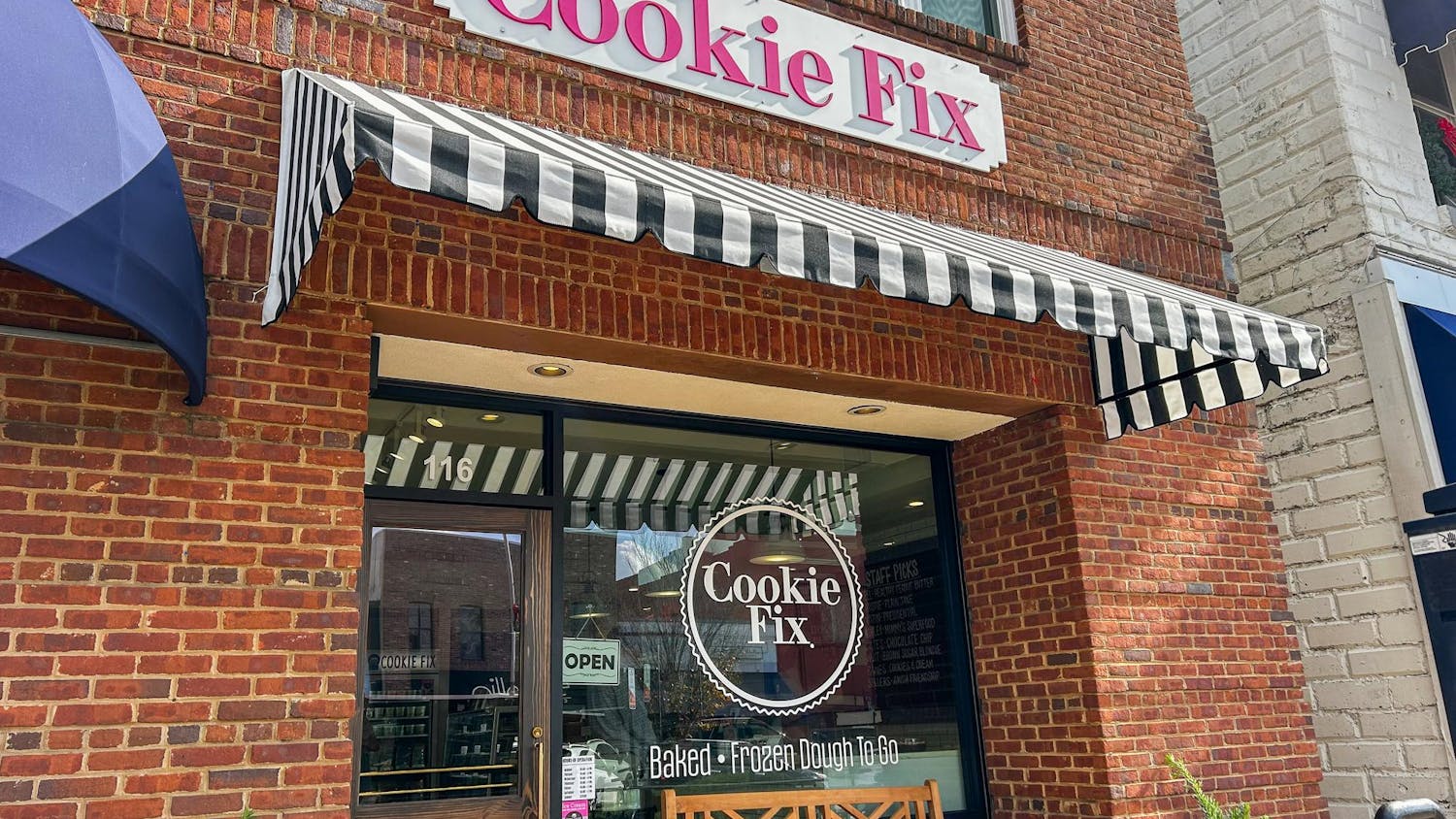 A brick storefront with a sign reading "Cookie Fix," featuring striped awnings, a glass door, and a wooden bench outside.