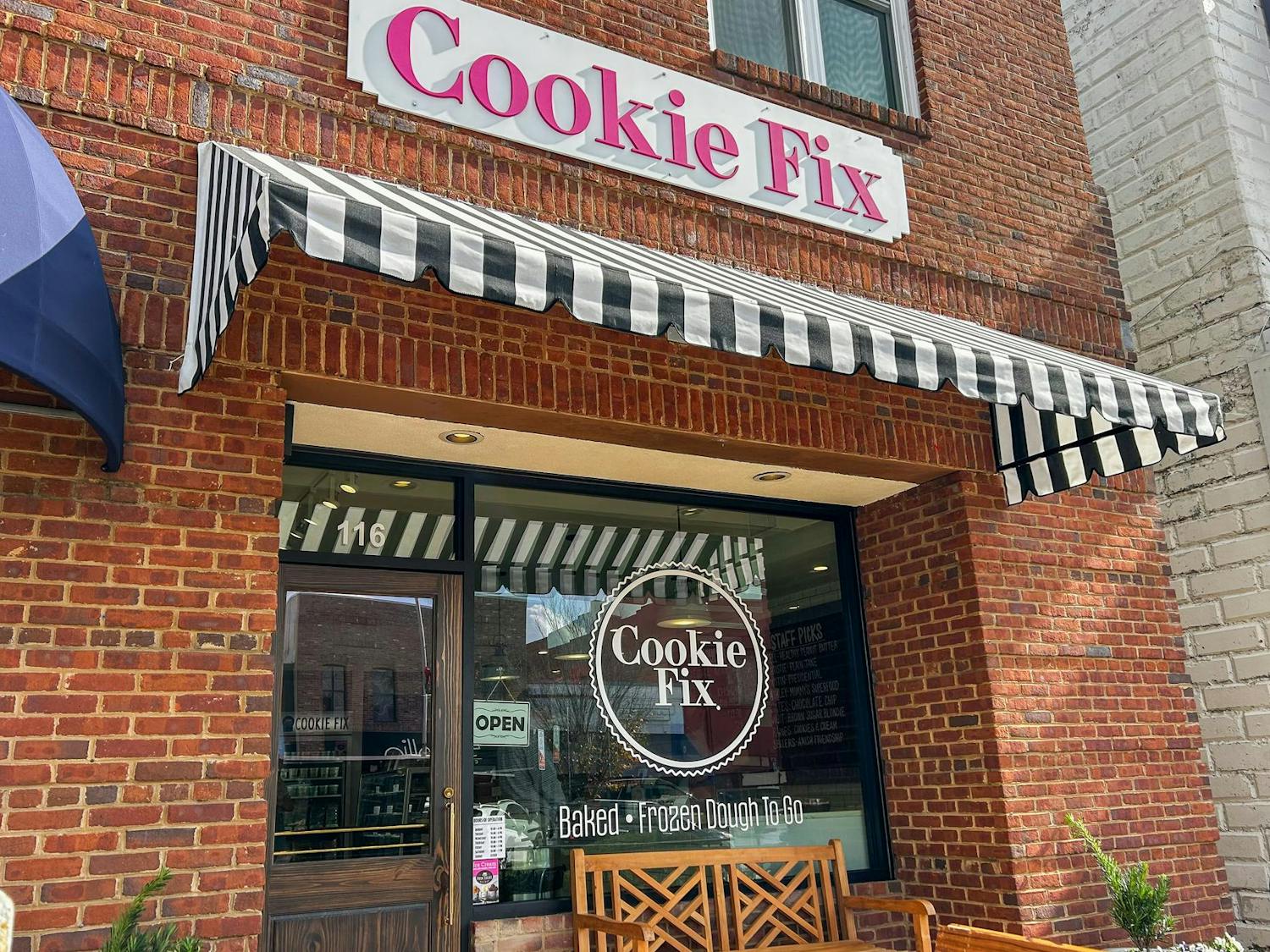 A brick storefront with a sign reading "Cookie Fix," featuring striped awnings, a glass door, and a wooden bench outside.