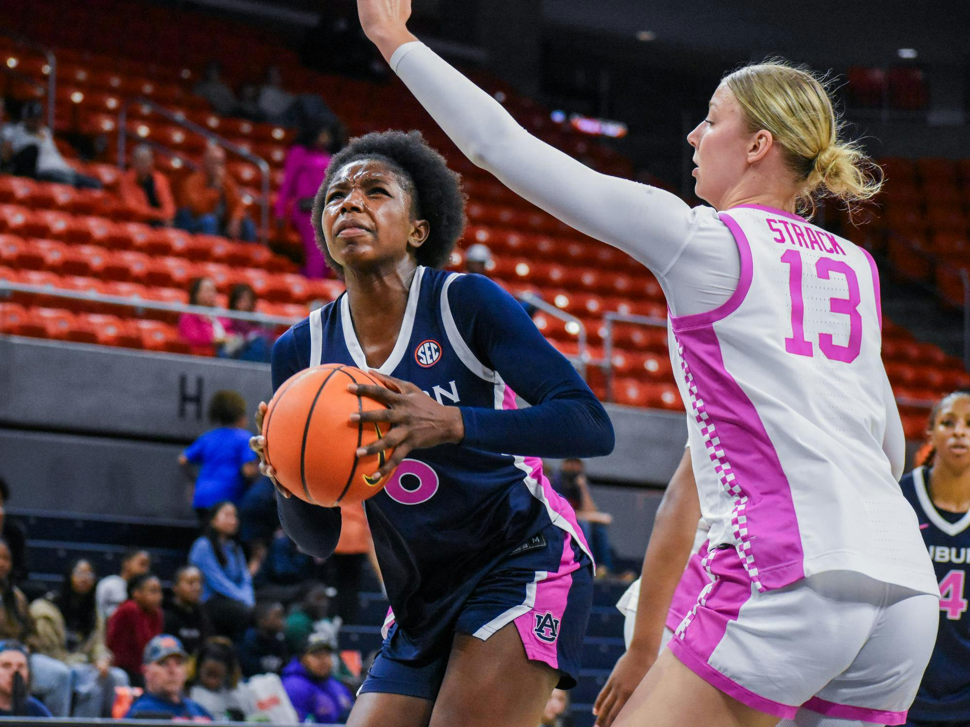 A player dressed in blue prepares to shoot a basketball while being defended by a player in white with pink accents.