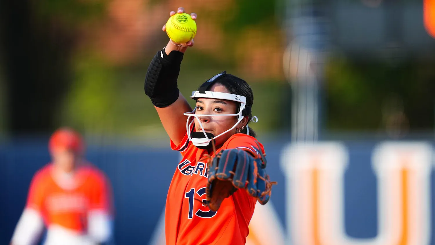 A young woman in a bright orange jersey is poised to pitch a yellow softball, with a blurred teammate visible behind her.