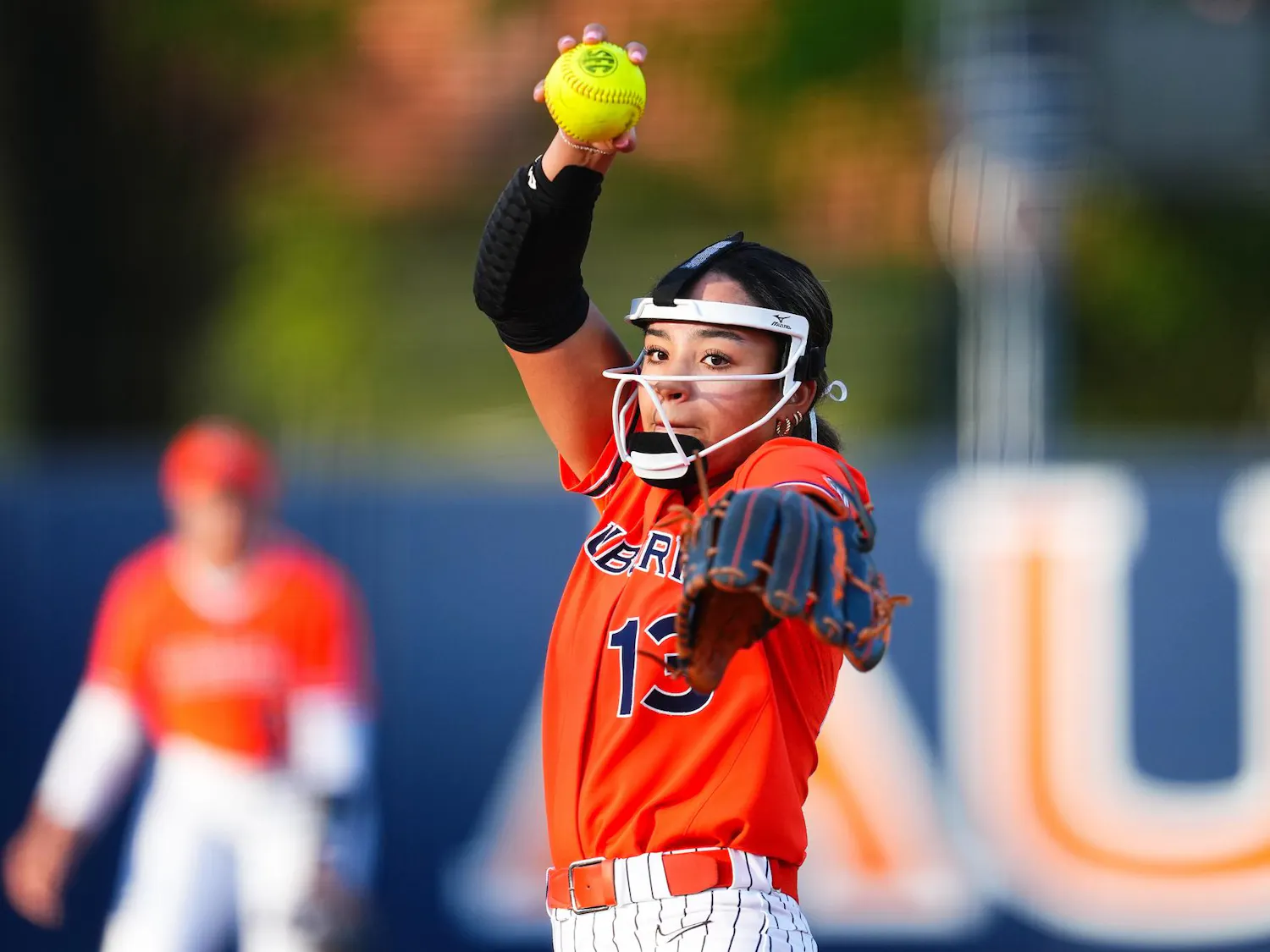 A young woman in a bright orange jersey is poised to pitch a yellow softball, with a blurred teammate visible behind her.