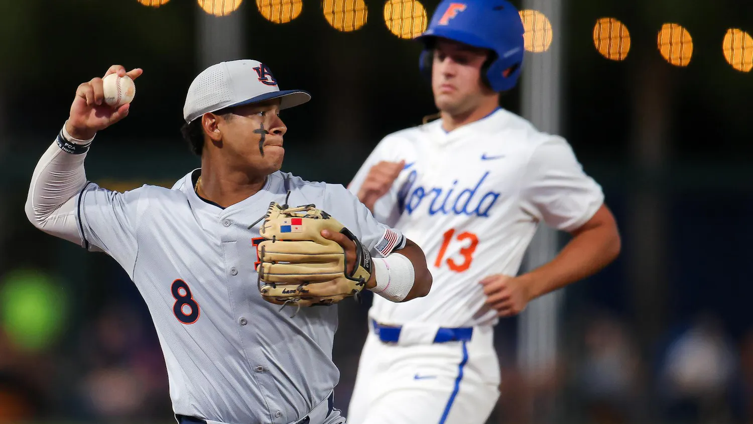 A baseball player in gray throws a ball while a runner in white approaches from behind, with lights glowing in the background.