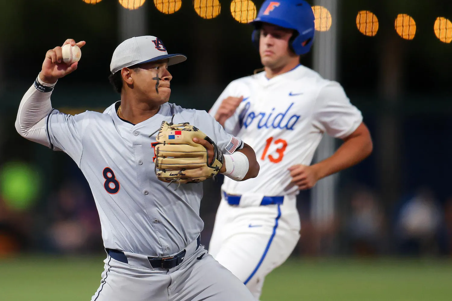 A baseball player in gray throws a ball while a runner in white approaches from behind, with lights glowing in the background.