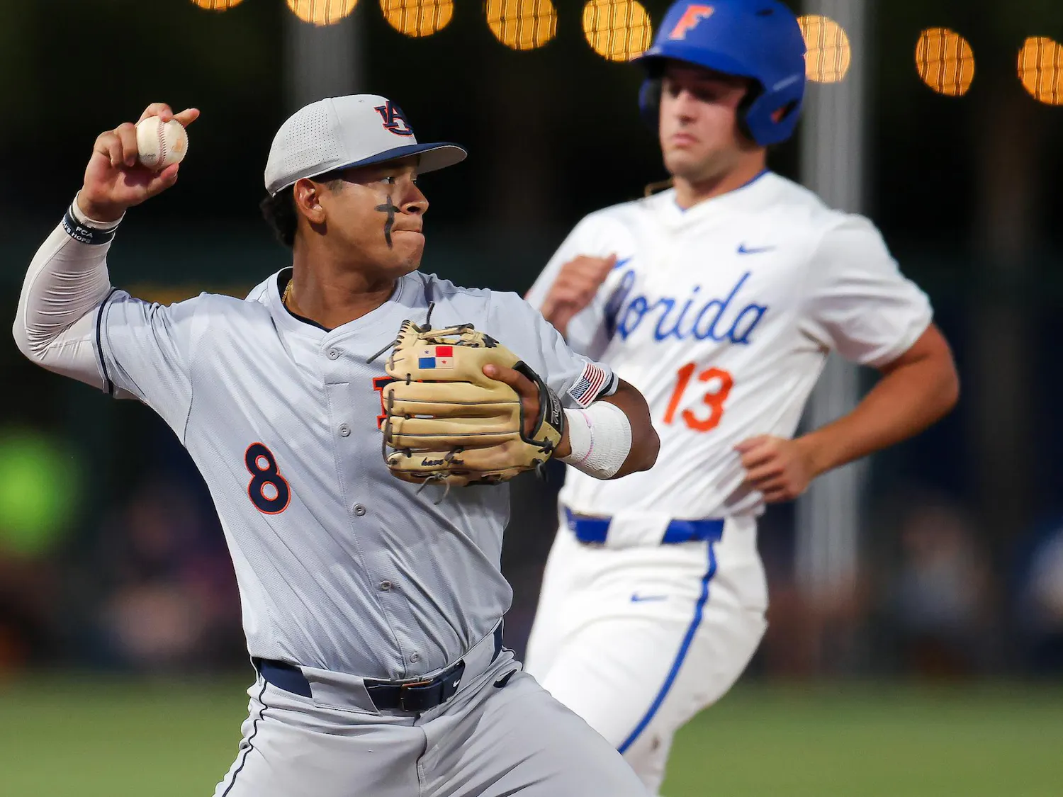 A baseball player in gray throws a ball while a runner in white approaches from behind, with lights glowing in the background.