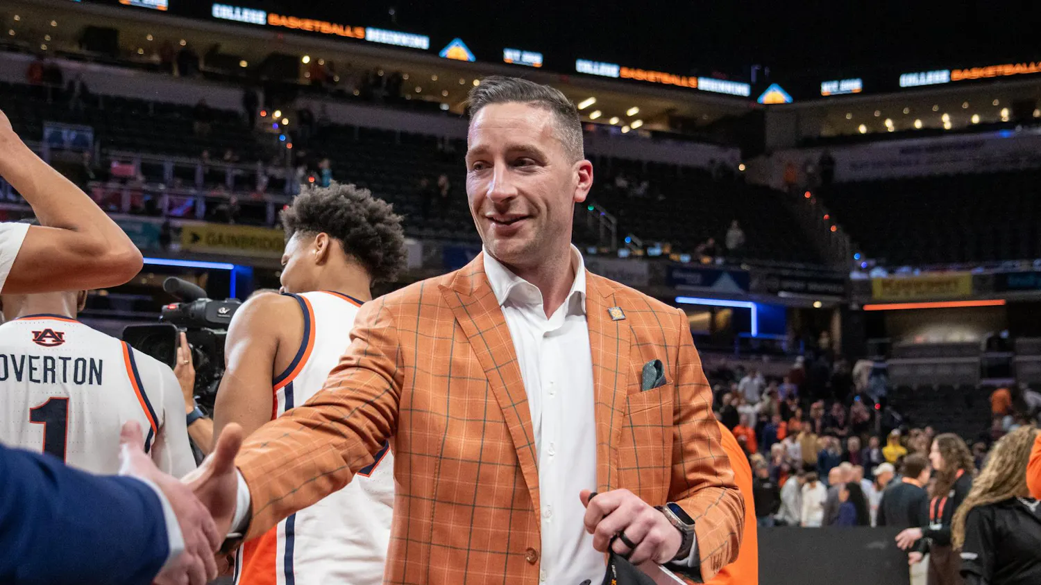 A man in a plaid orange suit shakes hands with another person, while basketball players and a crowd are in the background.