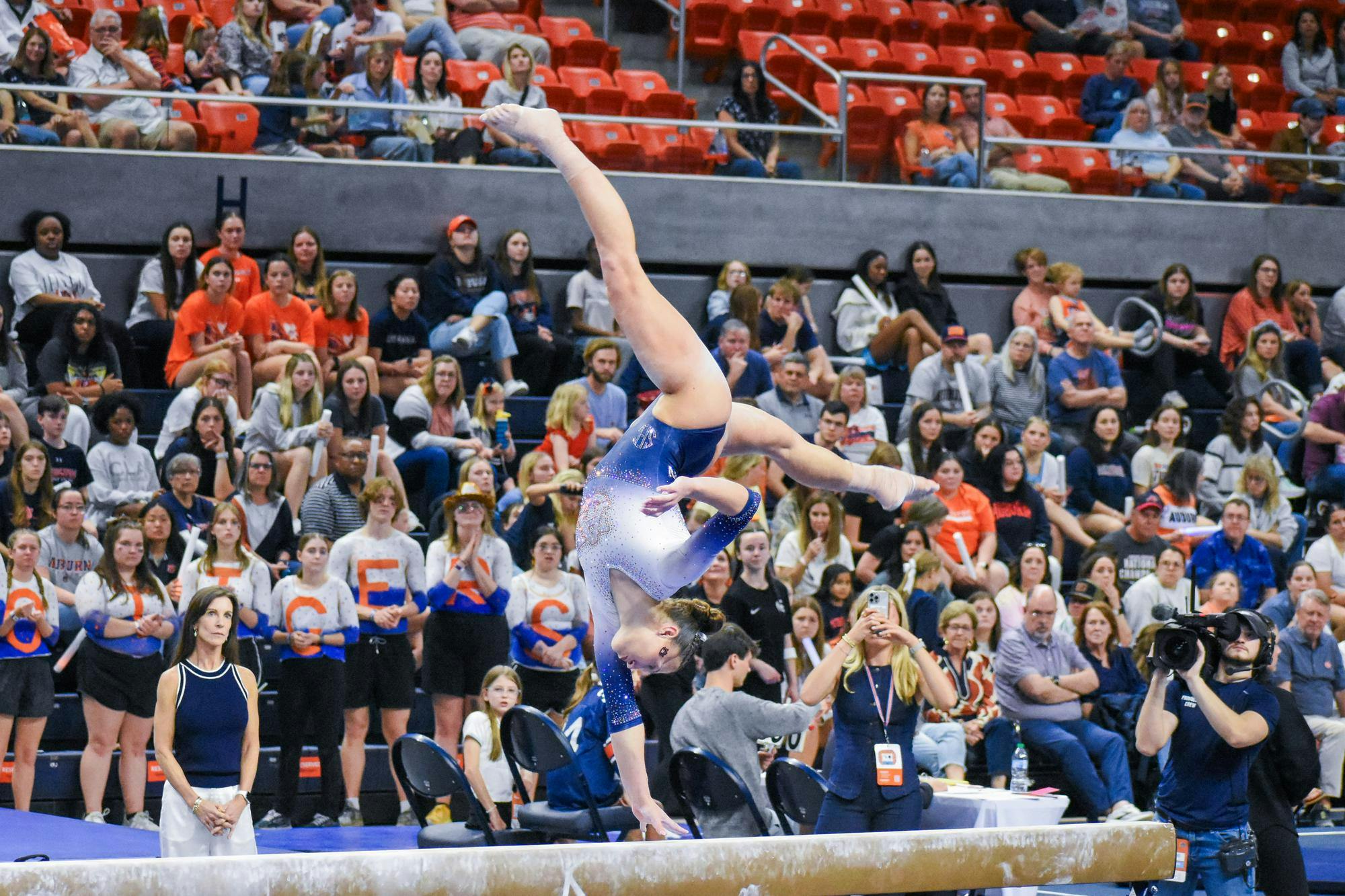 A gymnast performs an intricate move on a balance beam, surrounded by an audience of cheering fans.