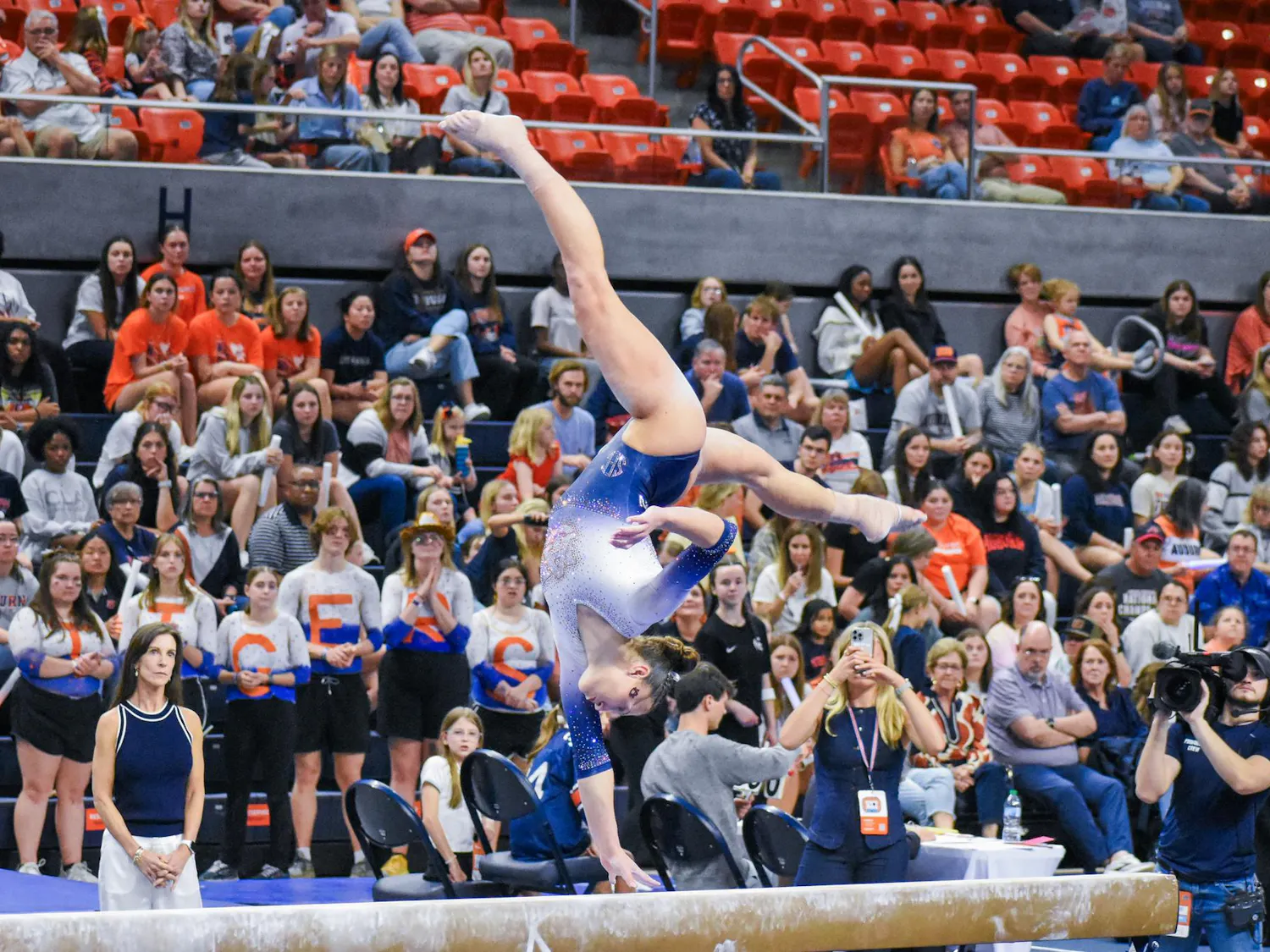 A gymnast performs an intricate move on a balance beam, surrounded by an audience of cheering fans.