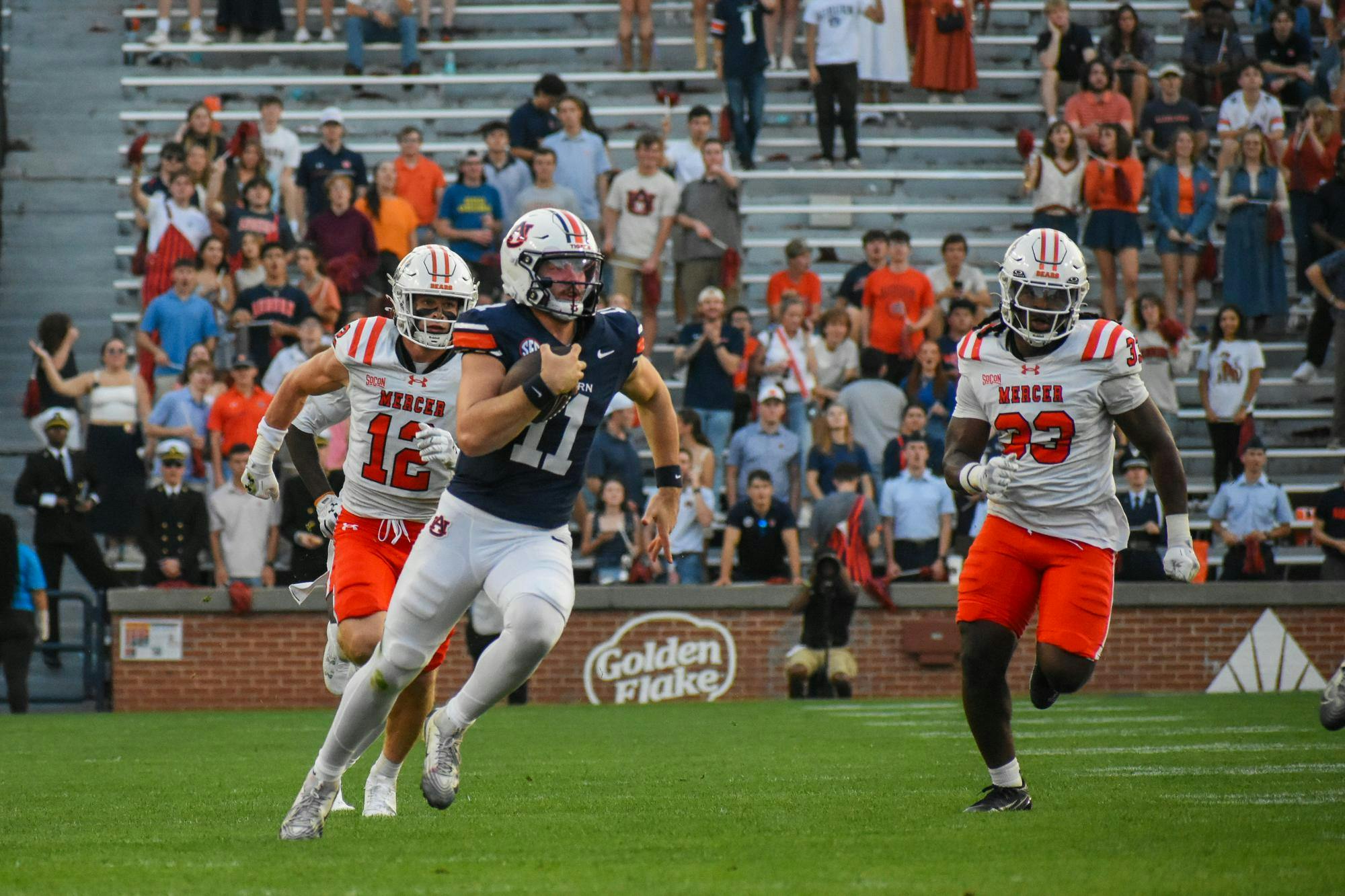 A football player in navy is running toward the end zone while being pursued by opponents in white and orange uniforms.