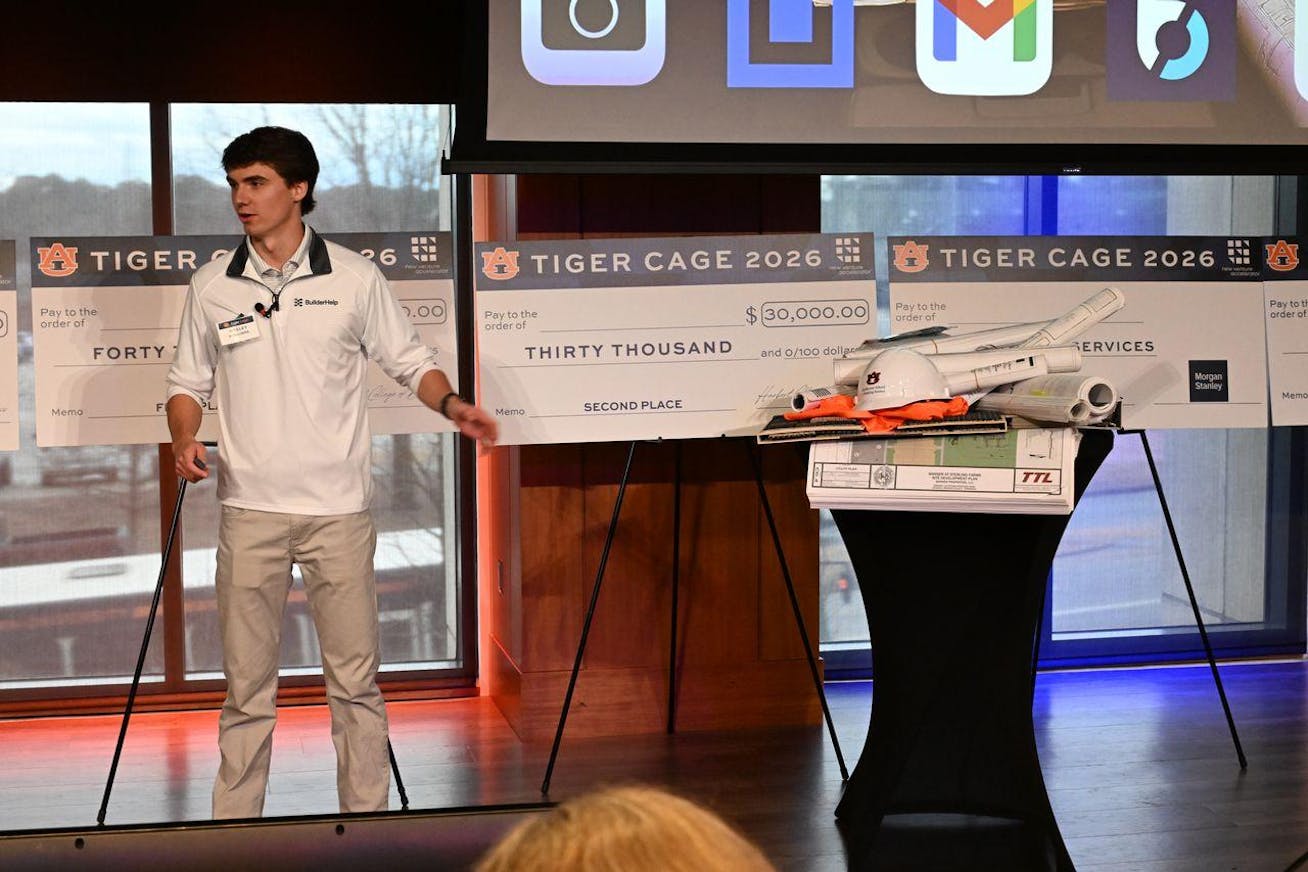 A young man in a white shirt stands at a podium beside large novelty checks and a model comprising blueprints and construction materials.