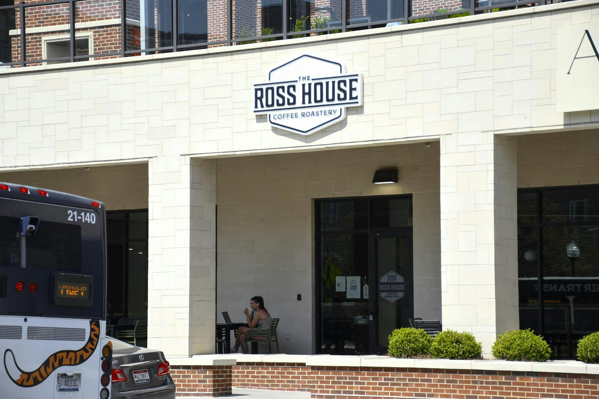 A woman sits at a table with a laptop outside "The Ross House Coffee Roastery," next to parked vehicles.