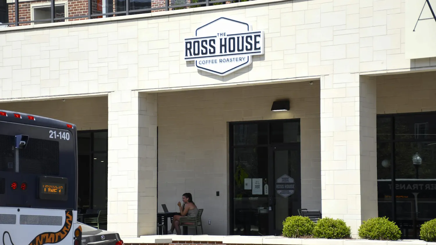 A woman sits at a table with a laptop outside "The Ross House Coffee Roastery," next to parked vehicles.