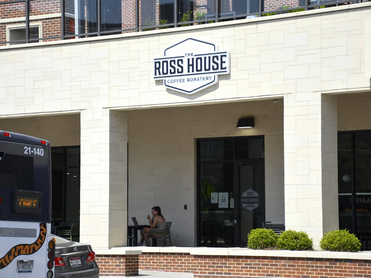 A woman sits at a table with a laptop outside "The Ross House Coffee Roastery," next to parked vehicles.