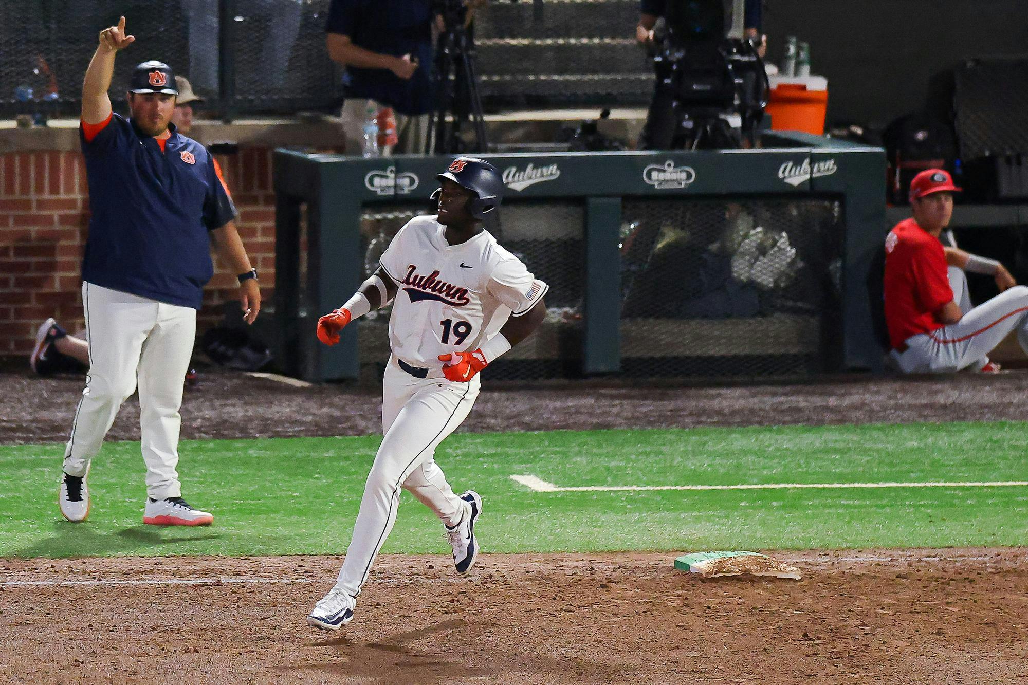 A baseball player in a white uniform with "Auburn" printed on it confidently runs toward first base while a coach signals from the sidelines.