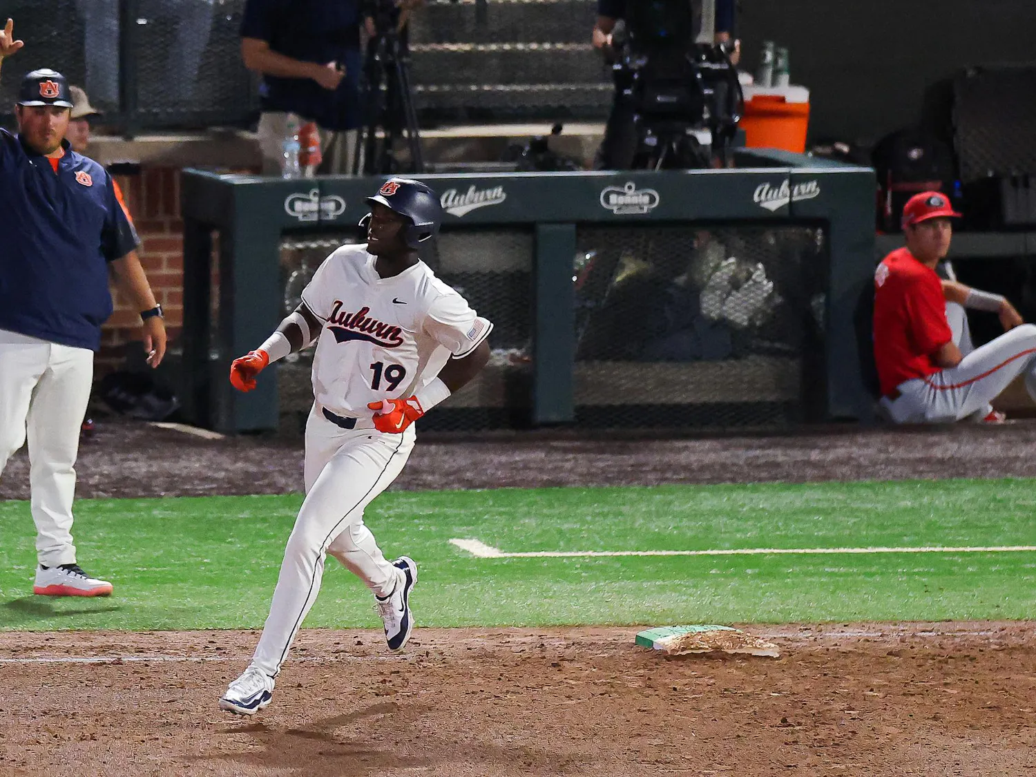 A baseball player in a white uniform with "Auburn" printed on it confidently runs toward first base while a coach signals from the sidelines.