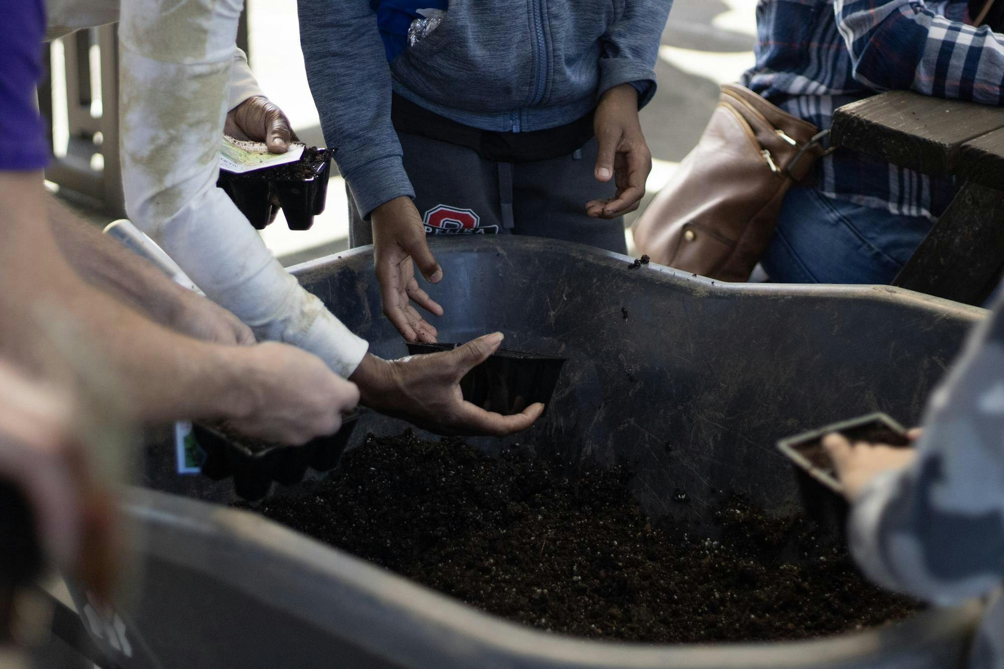 Hands of several people reach toward a container filled with soil, holding planting trays and preparing to fill them.