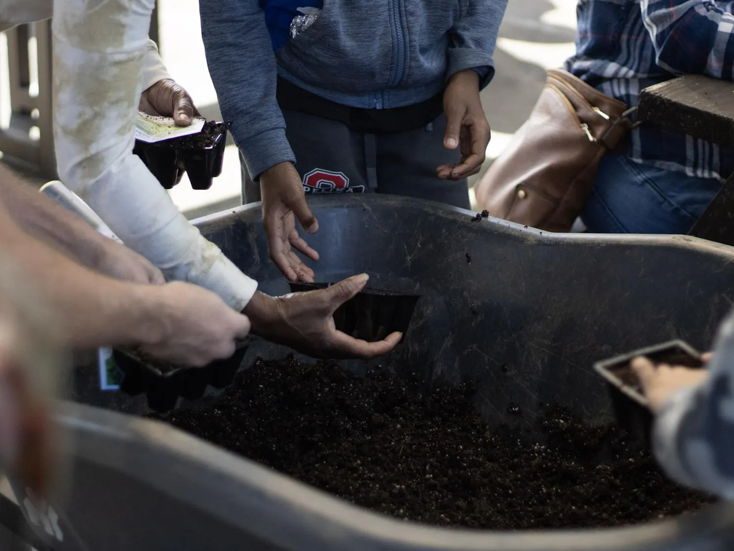 Hands of several people reach toward a container filled with soil, holding planting trays and preparing to fill them.