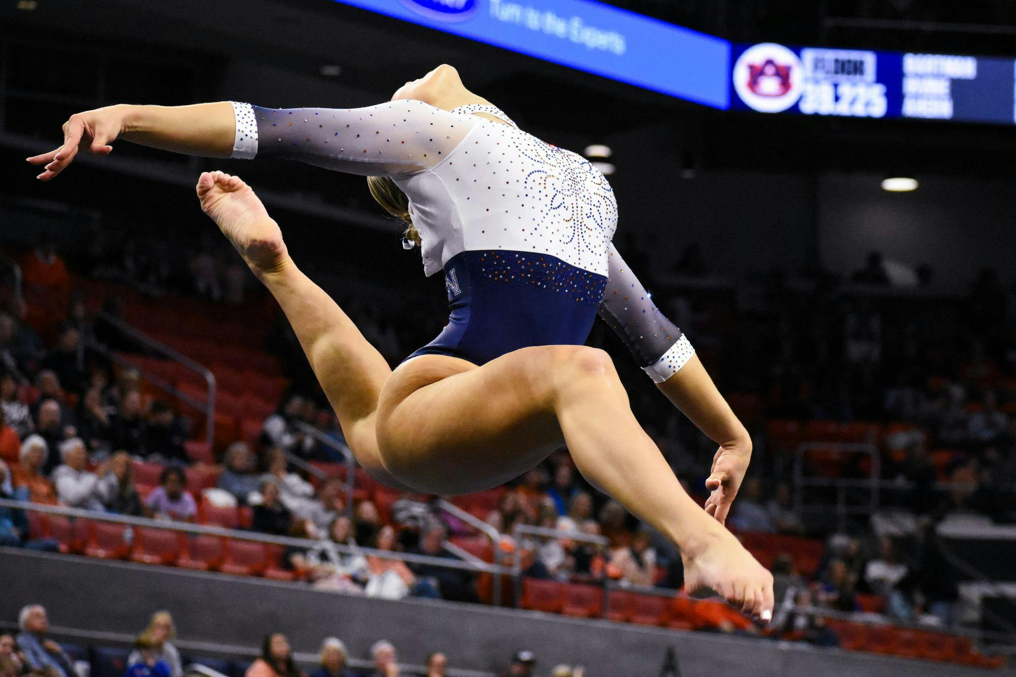 A gymnast performs an aerial leap, showcasing athleticism in a sparkly leotard above a crowd in a gymnasium.