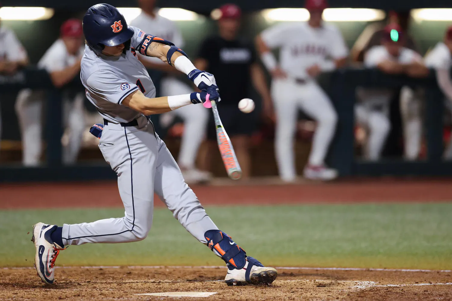 A baseball player in gray athletic gear swings a bat at a flying baseball, surrounded by teammates in the background.