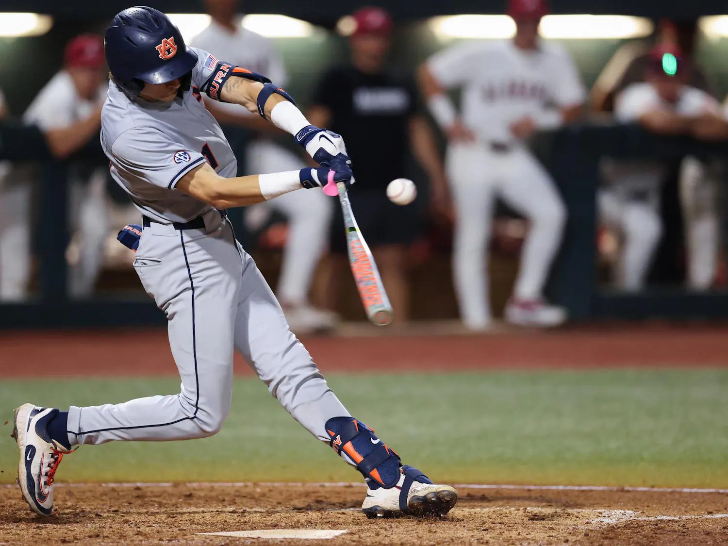A baseball player in gray athletic gear swings a bat at a flying baseball, surrounded by teammates in the background.