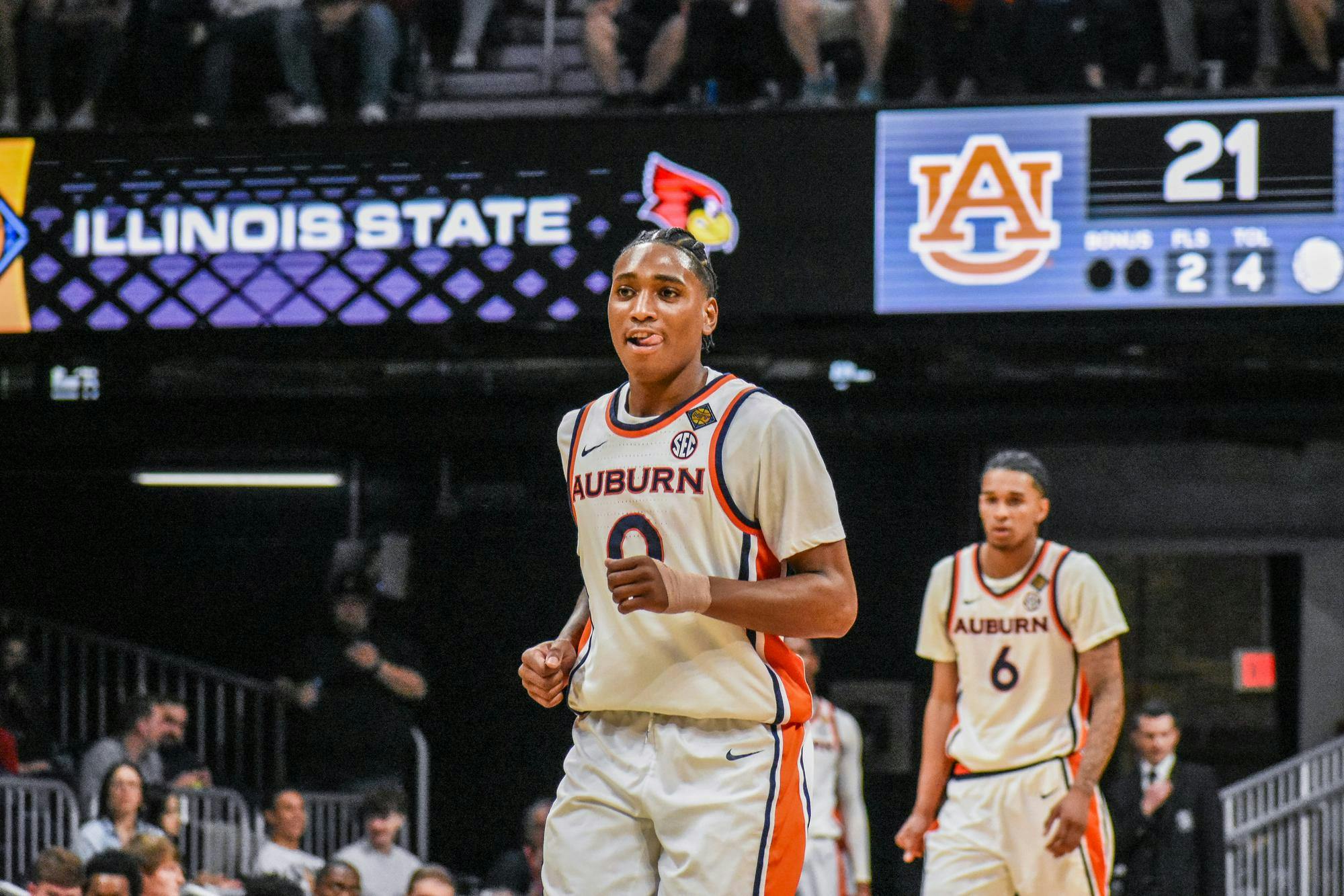 A basketball player wearing an Auburn jersey smiles and gestures on the court, with a scoreboard showing a game score in the background.