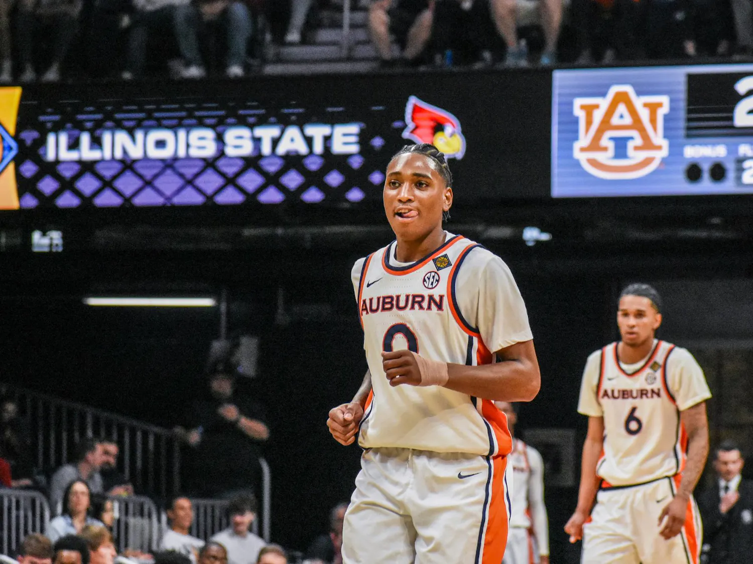 A basketball player wearing an Auburn jersey smiles and gestures on the court, with a scoreboard showing a game score in the background.