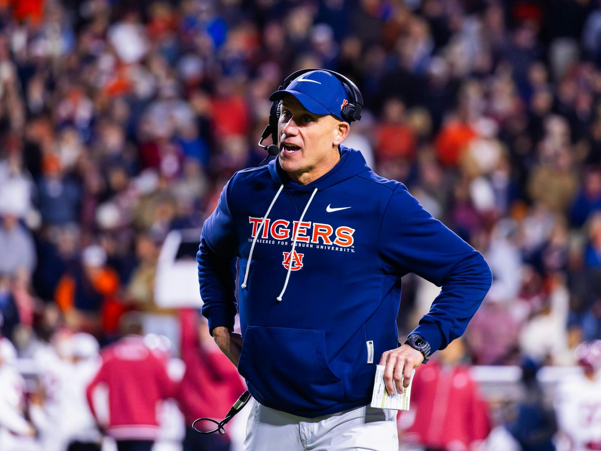 A man in a blue sweatshirt with "TIGERS" written on it stands on the sidelines of a football game, holding papers and wearing a headset.